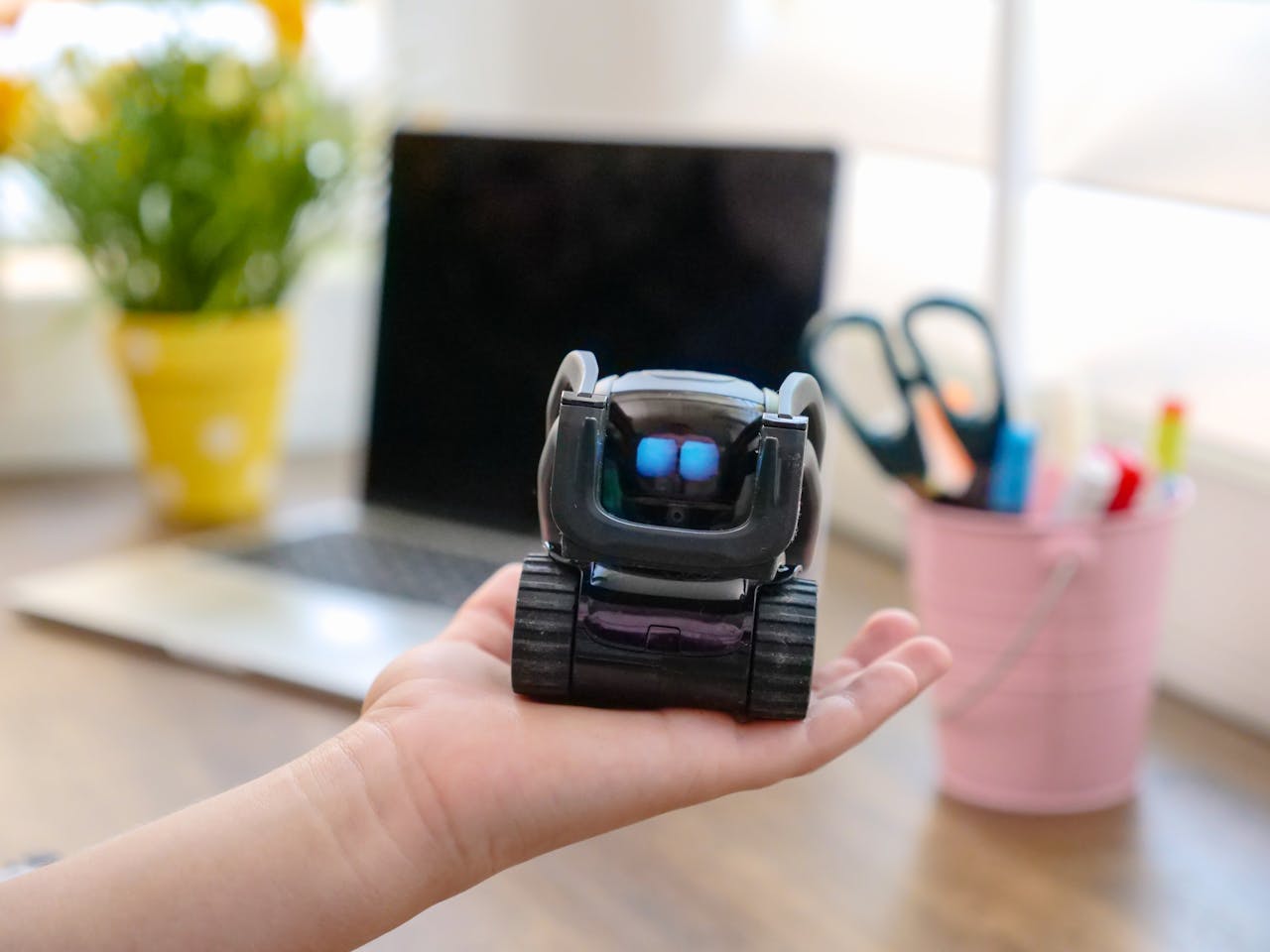 A modern toy robot held in a hand with an office desk in the background, showcasing innovation.